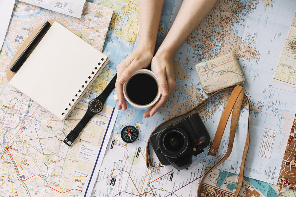 hand holding coffee cup map surrounded with spiral notepad compass wristwatch camera strap 1