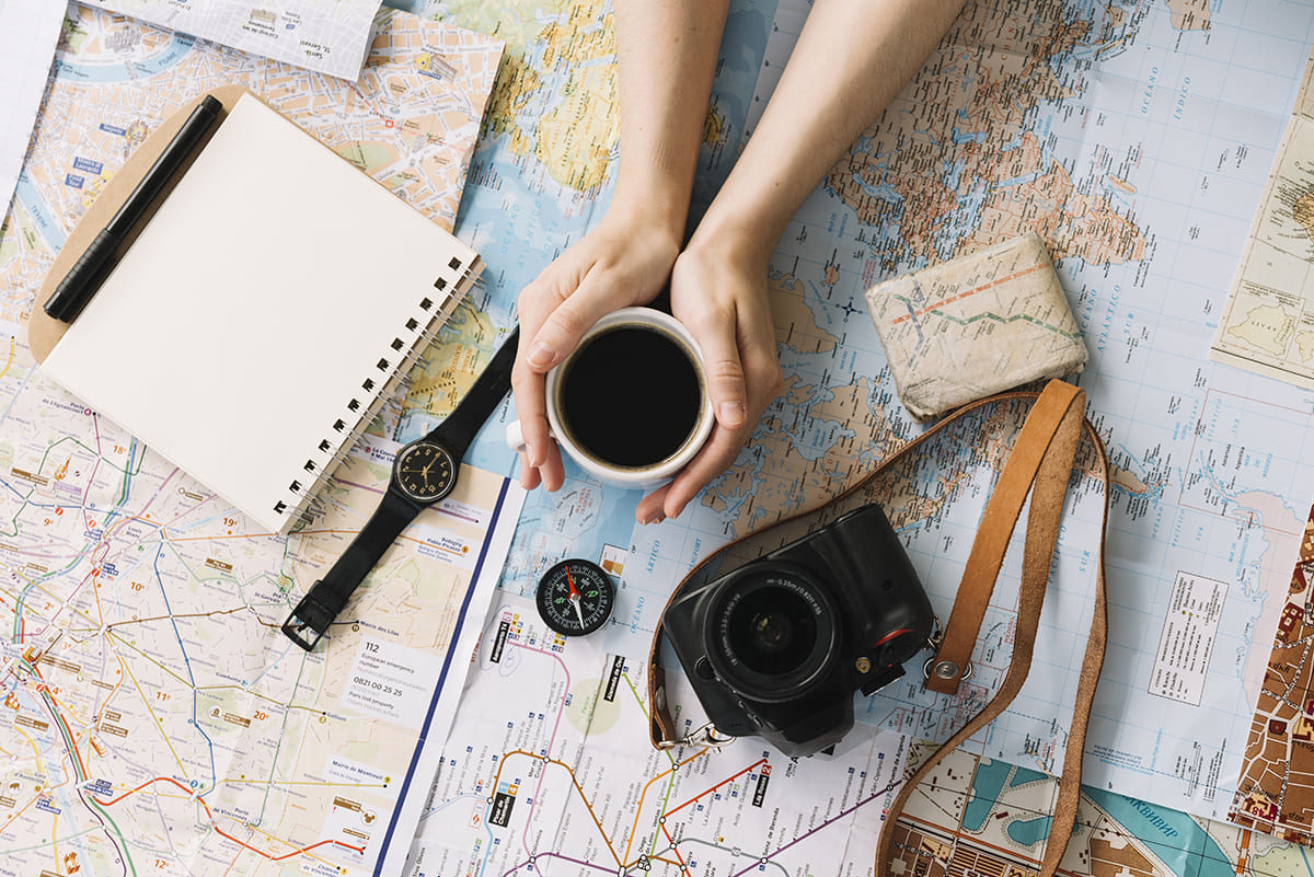 hand holding coffee cup map surrounded with spiral notepad compass wristwatch camera strap 1