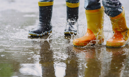 funny kids rain boots playing with paper ship by puddle