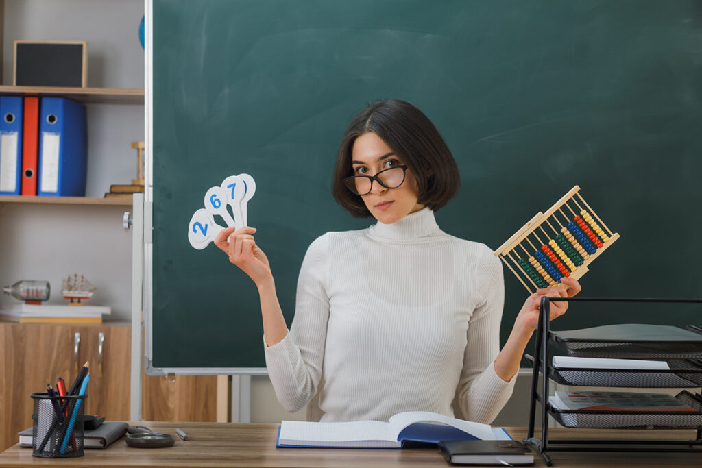 Белстат раскрыл, сколько зарабатывают учителя в Беларуси 2 strict young female teacher wearing glasses holding abacus with number fan sitting desk with school tools classroom