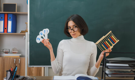 strict young female teacher wearing glasses holding abacus with number fan sitting desk with school tools classroom